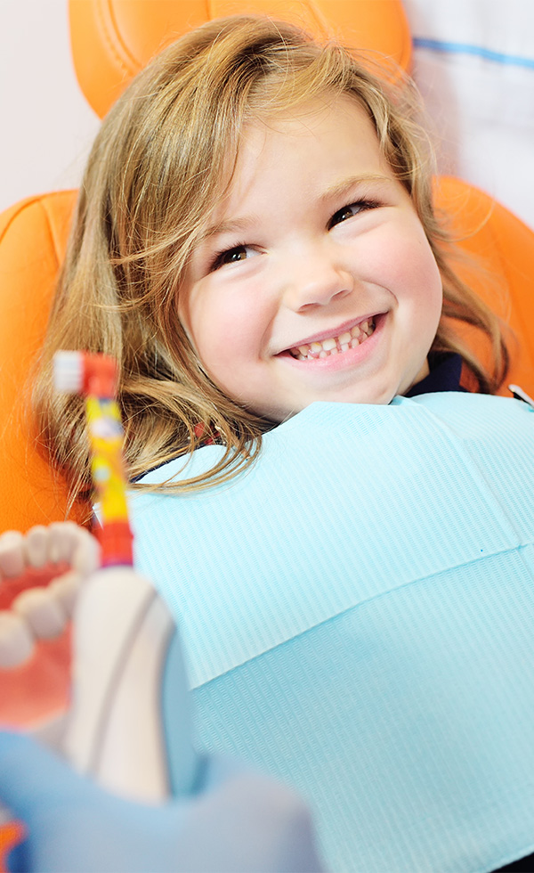 pediatric dentist shows how to properly brush her teeth to a little girl who is sitting in an orange dental chair and smiling